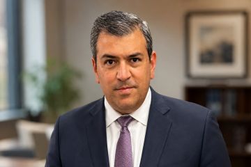 A man in a navy suit and purple tie smiles for a portrait in front of a blurred office background.