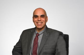 Mark Rusin, wearing a dark gray suit and maroon tie, smiles in front of a grayish white background